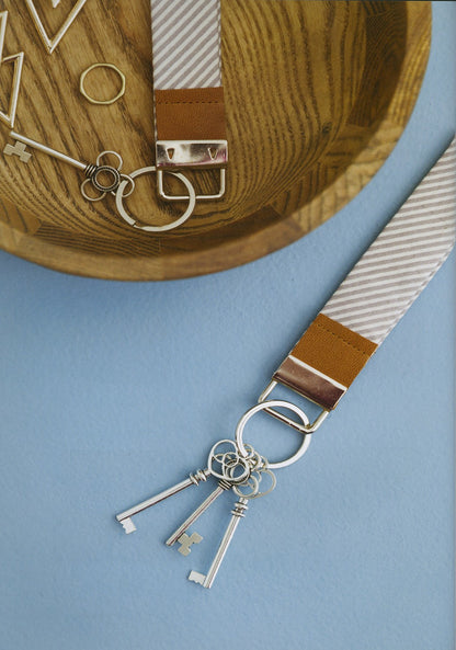 A close-up of a keychain accessory laid on a light blue surface, featuring a striped fabric strap with tan leather trim, metal key rings, and three silver key charms. A wooden bowl with additional key rings and decorative keys is partially visible at the top.
Photo by See Kate Sew [additional_image_link]