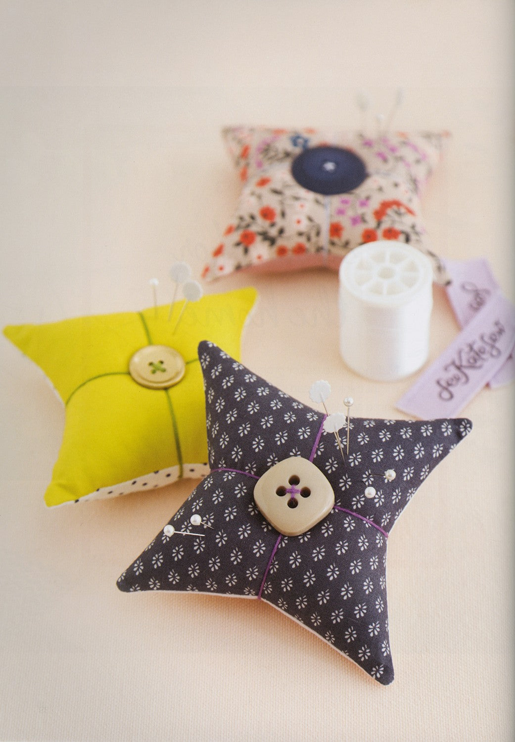 Three decorative star-shaped sewing pincushions arranged on a light beige surface, alongside a white spool of thread and a small lavender tag. One pincushion is bright yellow, one is dark gray with a small floral pattern and a cream button, and one has a pink floral print with a dark blue button.
Photo by See Kate Sew [additional_image_link]