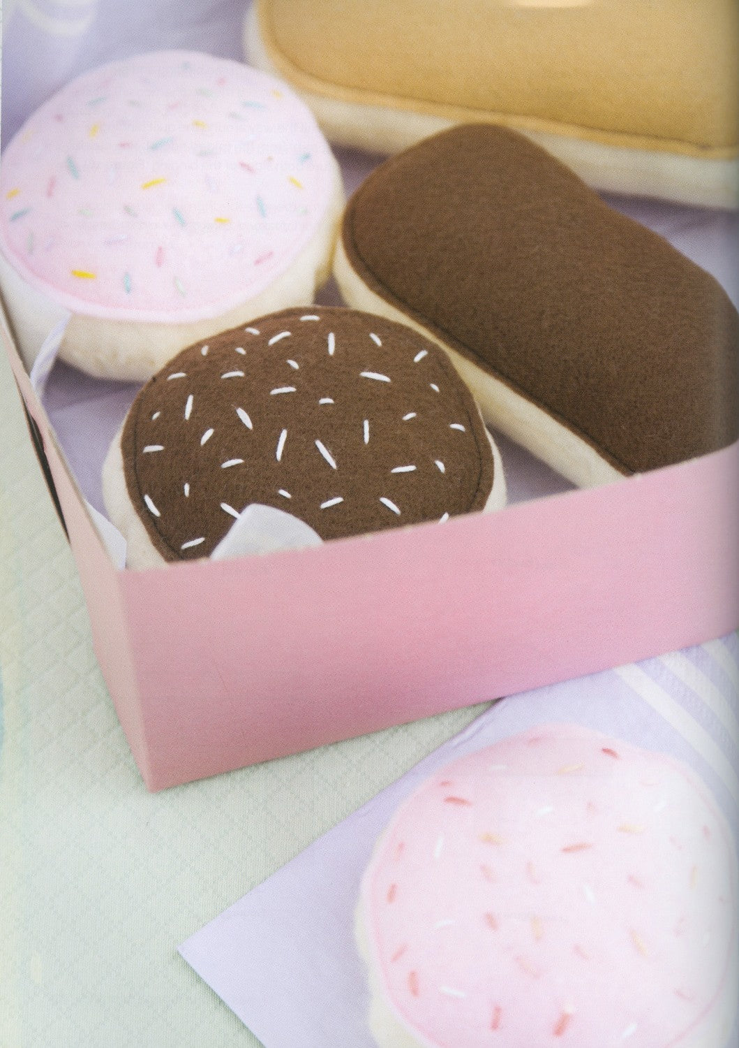 A pink gift box containing plush donut-shaped toys, including one with brown frosting and sprinkles and one with pink frosting and sprinkles, on a pale surface.
Photo by See Kate Sew [additional_image_link]