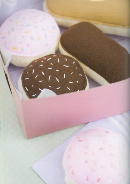 A pink gift box containing plush donut-shaped toys, including one with brown frosting and sprinkles and one with pink frosting and sprinkles, on a pale surface.
Photo by See Kate Sew [additional_image_link]