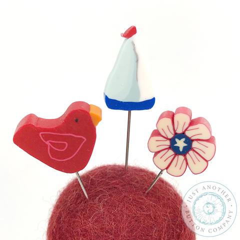 A pincushion kit from Just Another Button Company, featuring a red felted wool ball, a wooden spool, and two seaworthy ribbons.
Pins include a sailboat, a flower and a red bird.
Photo by JustAnotherButtonCompany.com [additional_image_link] 