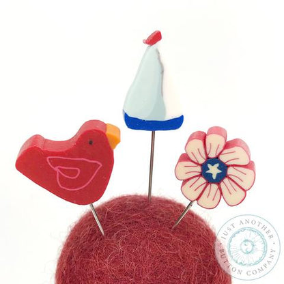 A pincushion kit from Just Another Button Company, featuring a red felted wool ball, a wooden spool, and two seaworthy ribbons.
Pins include a sailboat, a flower and a red bird.
Photo by JustAnotherButtonCompany.com [additional_image_link] 