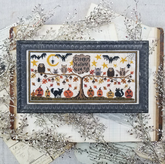A framed cross-stitch artwork featuring a Halloween tree with pumpkins, bats, owls, stars, and the text 'Sleep Hollow Leg 33', styled on top of vintage books and silver decorative branches.
Photo by HelloFromLizMathews.com [image_link]