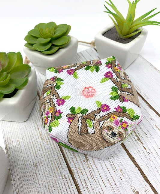 A small fabric-covered cross-stitch sampler box featuring a sloth and floral tree design, styled on a white wooden surface with small potted succulents in the background.
Photo by Tiny Modernist [image_link]