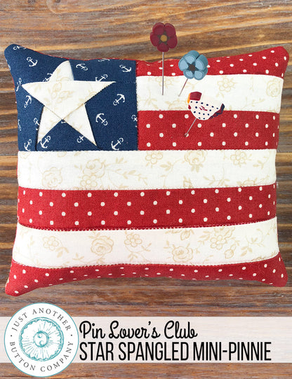A 'Star Spangled' mini pincushion by Just Another Button Company featuring a red, white, and blue color scheme with a star pattern, placed on a wooden surface with a 'Pin Lover's Club' label and branding.
Photo by JustAnotherButtonCompany.com [image_link]
