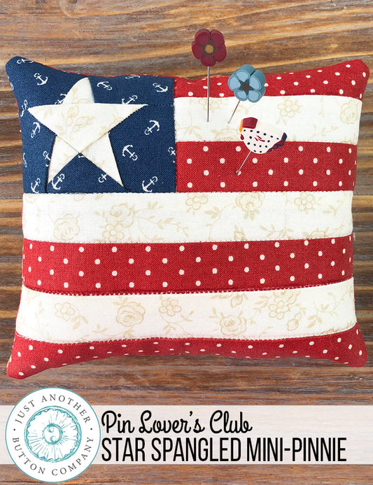 A 'Star Spangled' mini pincushion by Just Another Button Company featuring a red, white, and blue color scheme with a star pattern, placed on a wooden surface with a 'Pin Lover's Club' label and branding.
Photo by JustAnotherButtonCompany.com [image_link]