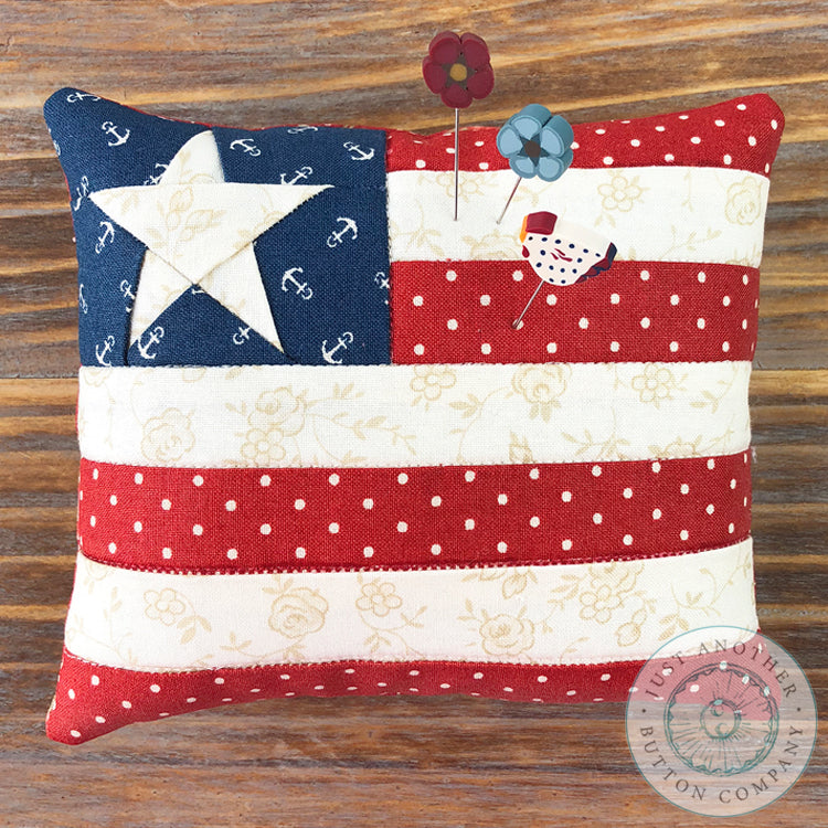 A 'Star Spangled' mini pincushion by Just Another Button Company featuring a red, white, and blue color scheme with a star pattern, placed on a wooden surface with a 'Pin Lover's Club' label and branding.
Pins included.
Photo by JustAnotherButtonCompany.com [additional_image_link]