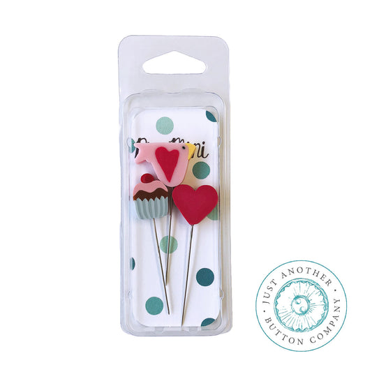 A packaged set of two metal sewing or button-needle tools with decorative tops: one pink bird and one cupcake with red hearts, shown in a clear blister pack on a white background. A circular 'Just Another Button Company' logo appears in the lower right.
Photo by JustAnotherButtonCompany.com [image_link]