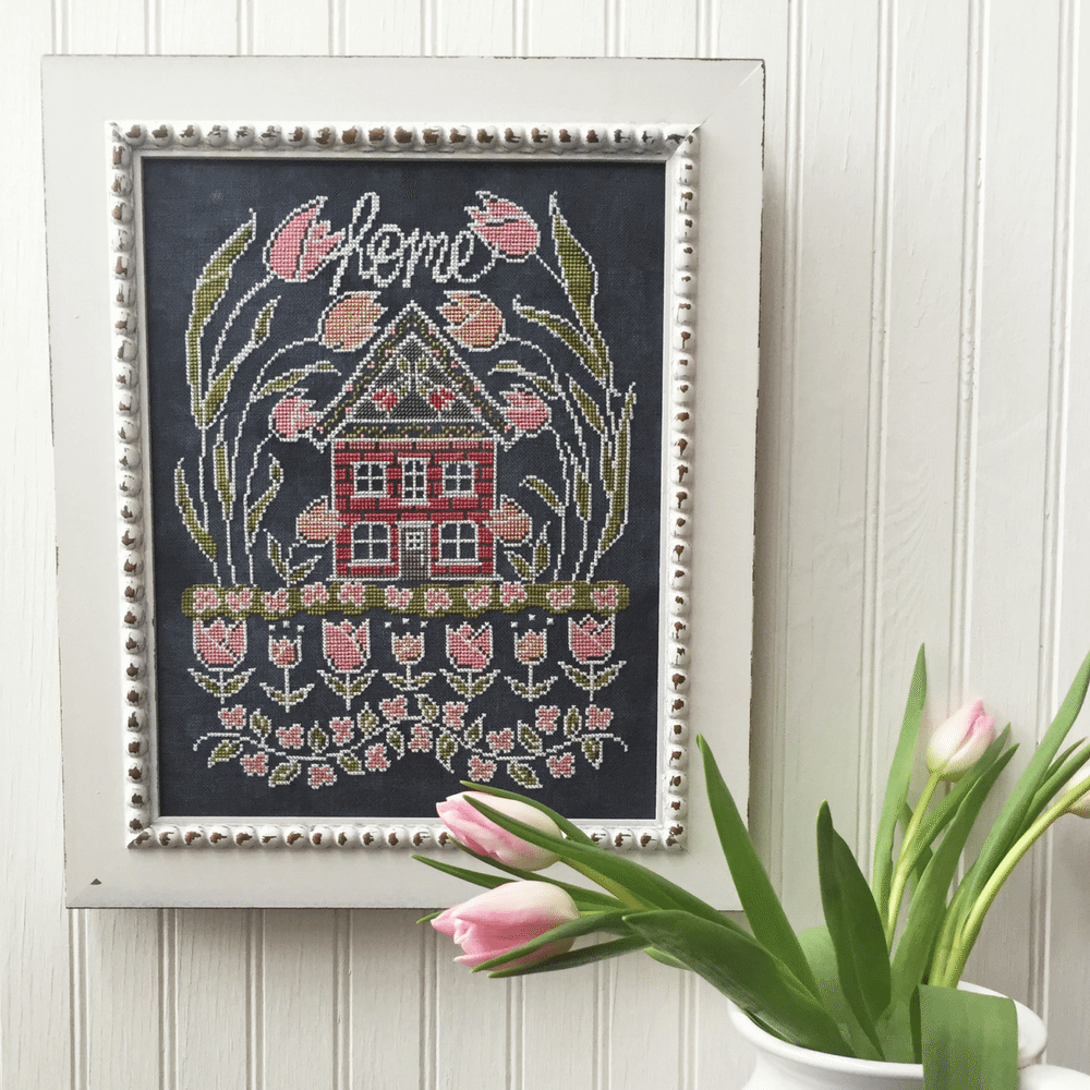 A framed cross-stitch artwork featuring a red house, pink tulips, and the word 'home' in white script on a dark background, styled on a white paneled wall with pink tulips in the foreground.
Photo by HandsOnDesign.biz [image_link]
