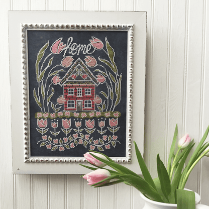 A framed cross-stitch artwork featuring a red house, pink tulips, and the word 'home' in white script on a dark background, styled on a white paneled wall with pink tulips in the foreground.
Photo by HandsOnDesign.biz [image_link]