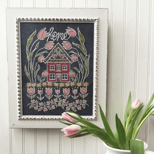 A framed cross-stitch artwork featuring a red house, pink tulips, and the word 'home' in white script on a dark background, styled on a white paneled wall with pink tulips in the foreground.
Photo by HandsOnDesign.biz [image_link]