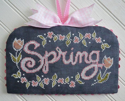 A handmade cross-stitch sampler featuring the word 'Spring' in pink cursive, surrounded by pink flowers and green leaves on a dark gray fabric background, finished with a pink gingham ribbon bow at the top.
Photo by HandsOnDesign.biz [additional_image_link]