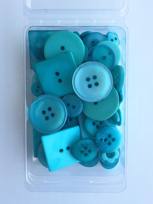A clear plastic storage container filled with assorted round and square buttons in shades of turquoise, teal, and light blue, photographed from above on a white background.
Photo by JustAnotherButtonCompany.com [image_link]