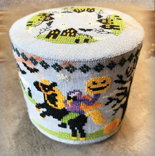 A cylindrical knitted pouf or ottoman featuring a colorful Halloween-themed needlepoint-style pattern with ghosts, pumpkins, bats, and haunted houses on a light gray base.
Photo by Tiny Modernist [image_link]