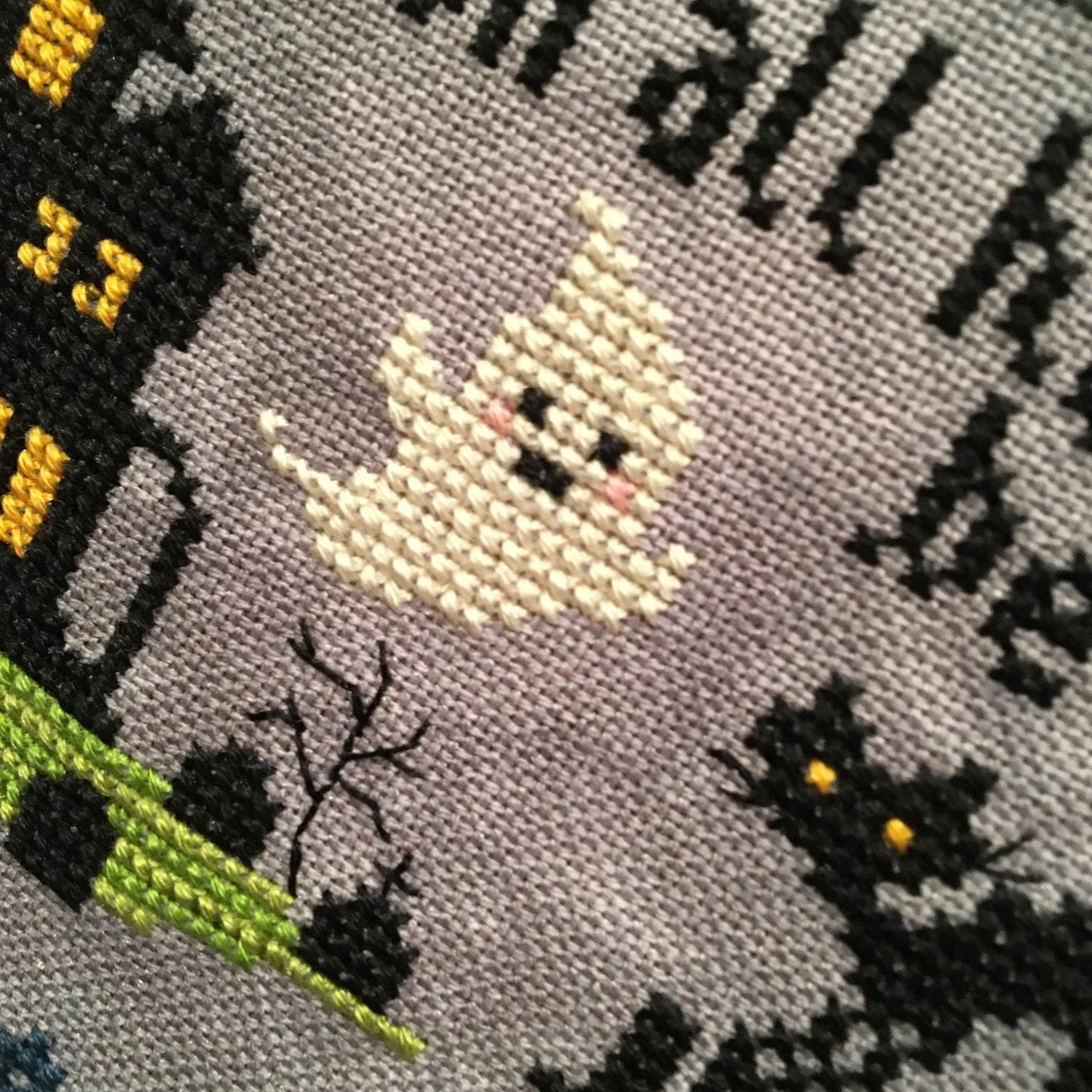 Close-up of a cross-stitch design featuring a smiling cream-colored ghost, a black haunted house with yellow windows, bare black branches, black cats with yellow eyes, and partial black text on a gray fabric background.
Photo by Tiny Modernist [additional_image_link]