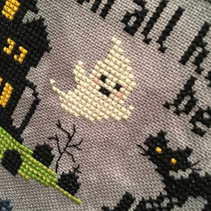 Close-up of a cross-stitch design featuring a smiling cream-colored ghost, a black haunted house with yellow windows, bare black branches, black cats with yellow eyes, and partial black text on a gray fabric background.
Photo by Tiny Modernist [additional_image_link]