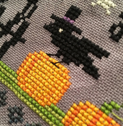 Close-up of a cross-stitch pattern on gray fabric, showing a black silhouette figure with a purple hatband above orange and yellow flowers with green stems.
Photo by Tiny Modernist [additional_image_link]