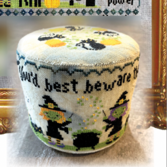 A round, upholstered pouf or ottoman with a cross-stitch style Halloween design. The light beige fabric features embroidered black text reading 'You'd best beware of' above cartoon witches, a cauldron, and a green cat, with black cat silhouettes on top.
Photo by Tiny Modernist [image_link]