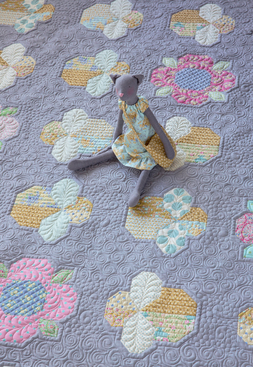 A small gray plush doll wearing a yellow floral dress sits on a lavender quilted blanket with embroidered flower motifs in pastel pink, cream, and yellow.
Photo by Tildafabrics.com [additional_image_link]