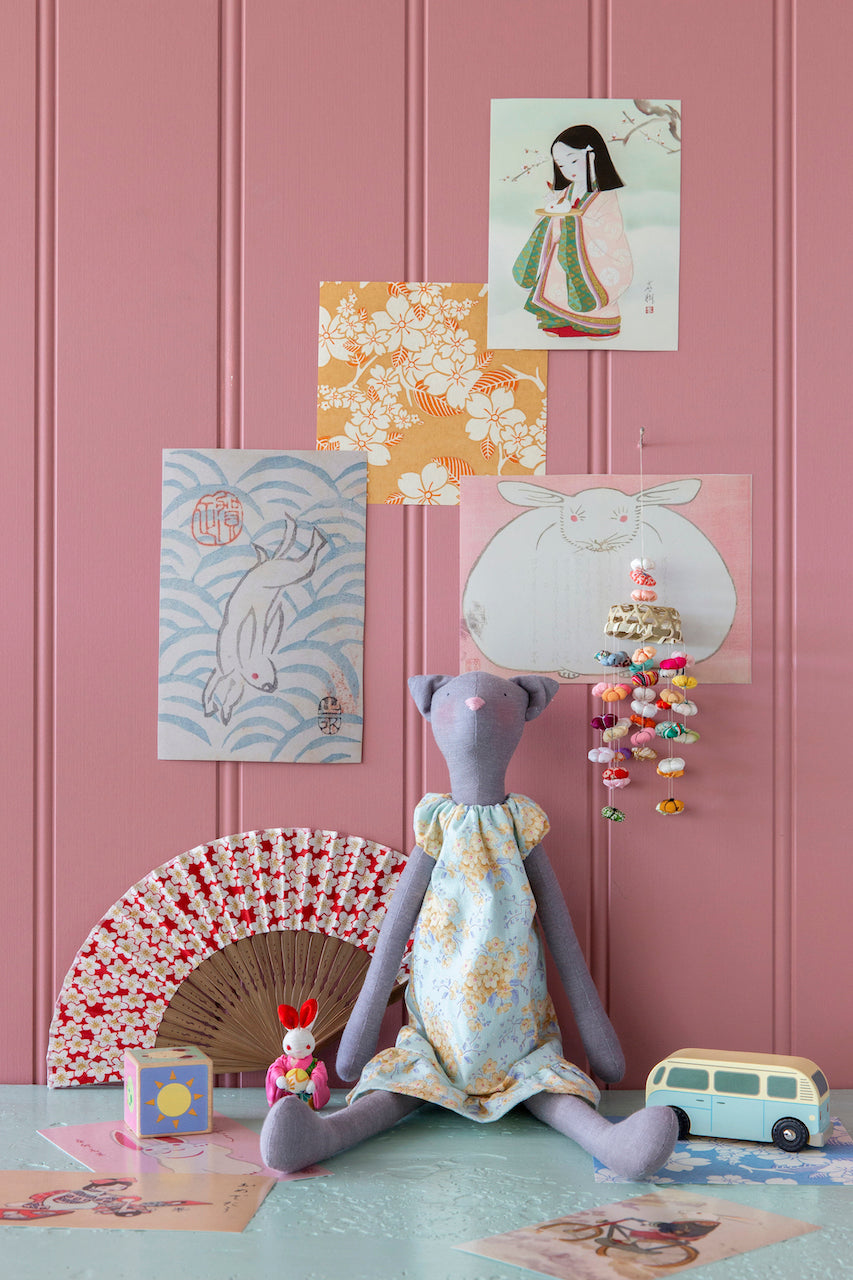 A styled interior scene with a lavender plush doll wearing a floral dress, seated against a pink paneled wall. Several art prints are pinned or hung on the wall, along with a folding fan, a small doll, a toy bus, and a hanging multicolored ornament.
Photo by Tildafabrics.com [additional_image_link]