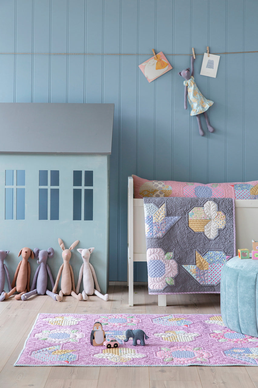 A styled children's bedroom corner featuring a lavender and gray patchwork quilt draped over a white bed, a matching lavender rug on the floor, a light blue house-shaped toy, and several plush bunny toys against a blue paneled wall.
Photo by Tildafabrics.com [additional_image_link]