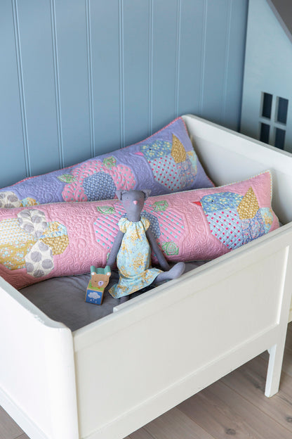 A white wooden toddler bed styled with two long patchwork quilted pillows in pastel pink, purple, and yellow, along with a small gray doll sitting on a gray mattress.
Photo by Tildafabrics.com [additional_image_link]