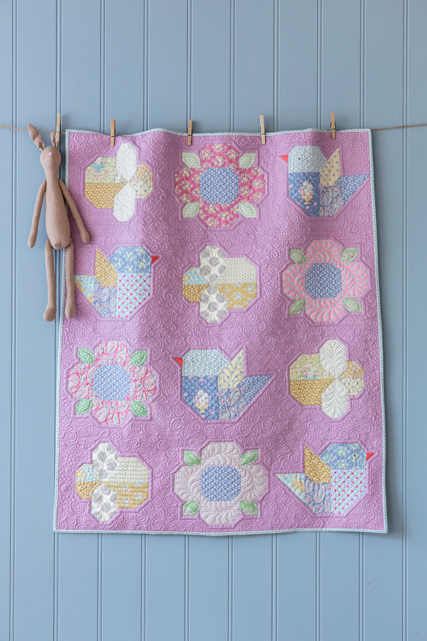 A handmade quilted textile featuring a lavender background and a grid of colorful bird and flower patchwork motifs, hanging from a string with wooden clothespins against a light blue paneled wall, with a small plush bunny hanging on the left.
Photo by Tildafabrics.com [additional_image_link]