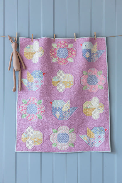 A handmade quilted textile featuring a lavender background and a grid of colorful bird and flower patchwork motifs, hanging from a string with wooden clothespins against a light blue paneled wall, with a small plush bunny hanging on the left.
Photo by Tildafabrics.com [additional_image_link]
