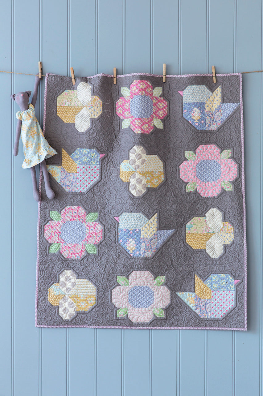 A handmade quilted textile featuring a grid of pastel bird and flower motifs on a gray background, hanging from a string with wooden clothespins against a light blue paneled wall. A small fabric doll is clipped to the left side.
Photo by Tildafabrics.com [additional_image_link]