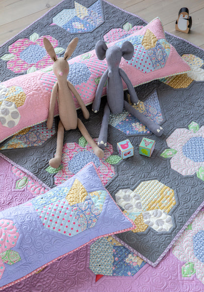 A styled flat-lay of a quilted textile set featuring a gray quilt with pastel floral and geometric patchwork, matching lavender pillows, two plush stuffed animals (a beige rabbit and a gray elephant), and small toy blocks on a light wood floor.
Photo by Tildafabrics.com [additional_image_link]