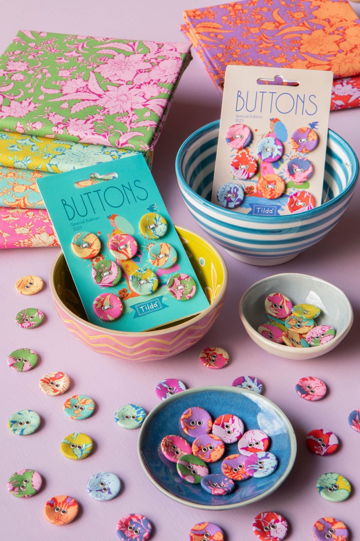 A styled flat-lay of colorful decorative buttons in various patterns, shown both on their retail backing cards and scattered on a pastel pink surface, with pastel bowls and patterned paper napkins in the background.
Photo by Tildafabrics.com [additional_image_link]