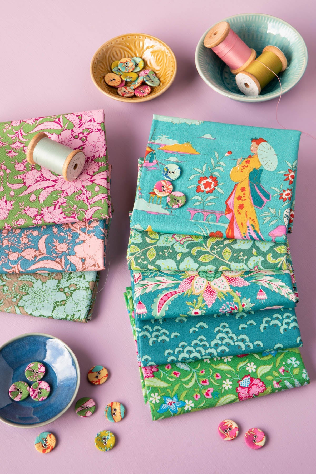 A styled flat-lay of multiple fabric bolts with colorful floral and traditional-inspired prints, arranged on a pink background with decorative spools of thread and buttons.
Photo by Tildafabrics.com [additional_image_link]