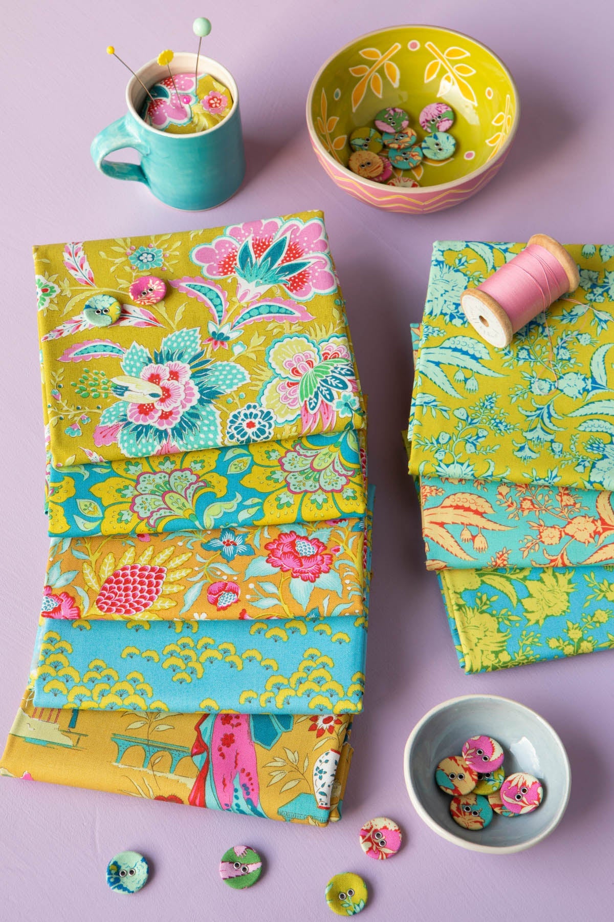 A styled flat-lay of colorful fabric bolts with floral and paisley patterns in yellow, teal, pink, and green, arranged with sewing props including buttons, a spool of pink thread, and a small bowl on a lavender background.
Photo by Tildafabrics.com [additional_image_link]