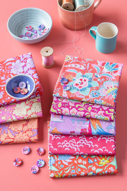 A styled flat-lay of multiple folded fabrics with colorful floral and patterned prints, arranged on a pink background with sewing-themed props including thread spools, buttons, a small bowl, and mugs.
Photo by Tildafabrics.com [additional_image_link]