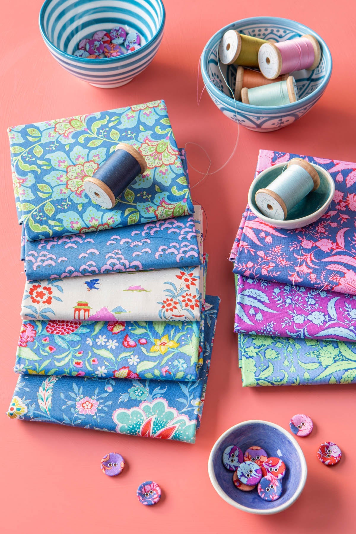 A styled flat-lay of multiple stacks of patterned fabric squares in various colors and prints, arranged with wooden spools of thread and small bowls of buttons on a solid coral pink background.
Photo by Tildafabrics.com [additional_image_link]
