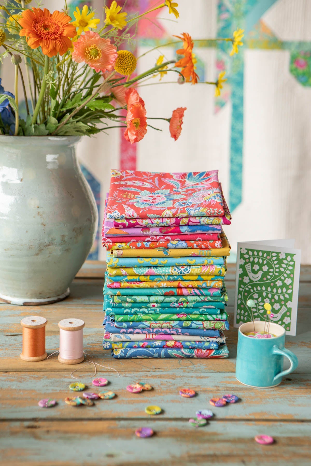 A stack of colorful patterned fabric squares sits on a rustic wooden table, styled with a large vase of orange and yellow flowers, two spools of thread, scattered buttons, and a turquoise mug.
Photo by Tildafabrics.com [additional_image_link]