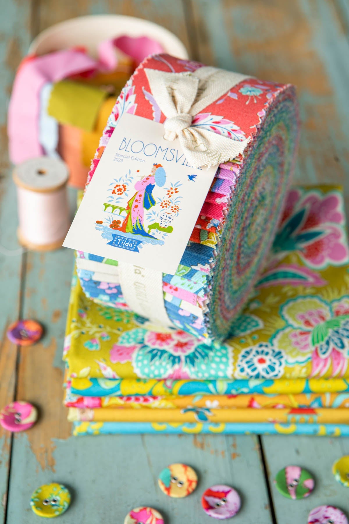 A bundle of colorful patterned fabric rolls tied with a cream ribbon and tag, displayed on top of a stack of matching fabric pieces on a rustic painted wood surface.
Photo by Tildafabrics.com [additional_image_link]