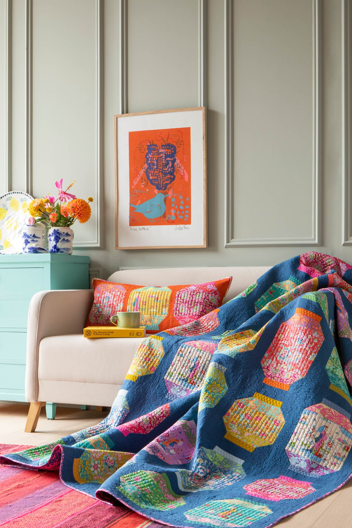 A colorful patterned throw quilt draped over a cream armchair in a styled room, with a matching patterned pillow, a framed art print on the wall, and a mint green side table with flowers.
Photo by Tildafabrics.com [additional_image_link]