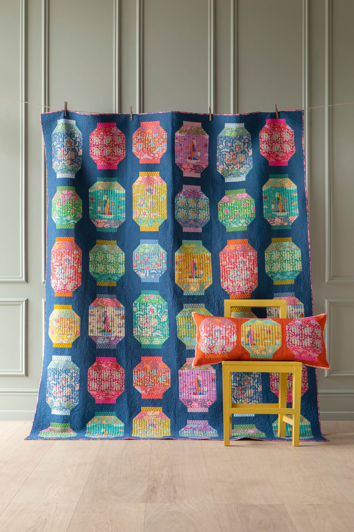 A colorful quilted blanket featuring a repeating pattern of octagonal lanterns in various bright colors on a navy blue background, draped over a yellow stool with matching decorative pillows in a room with paneled walls.
Photo by Tildafabrics.com [additional_image_link]