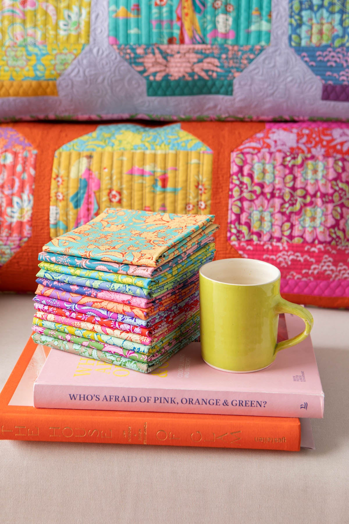 A stack of colorful patterned fabric squares sits on top of a pink book with an orange spine, next to a bright yellow-green ceramic mug. A quilted fabric piece with pink, orange, yellow, and purple pillows are draped in the background.
Photo by Tildafabrics.com [additional_image_link]