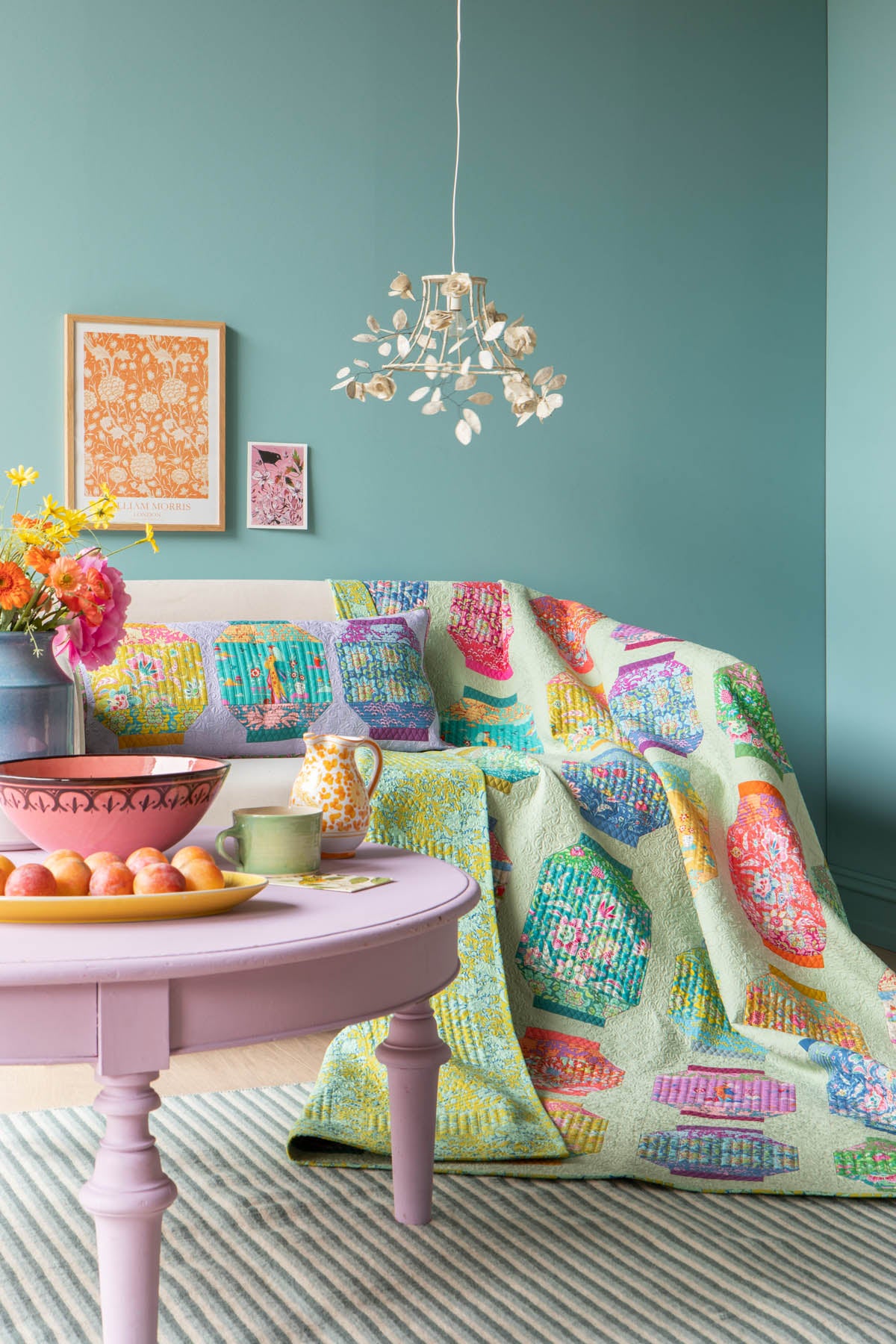 A colorful patterned quilt draped over a sofa in a styled living room, with matching patterned pillows, a pink round table, and a teal wall.
Photo by Tildafabrics.com [additional_image_link]