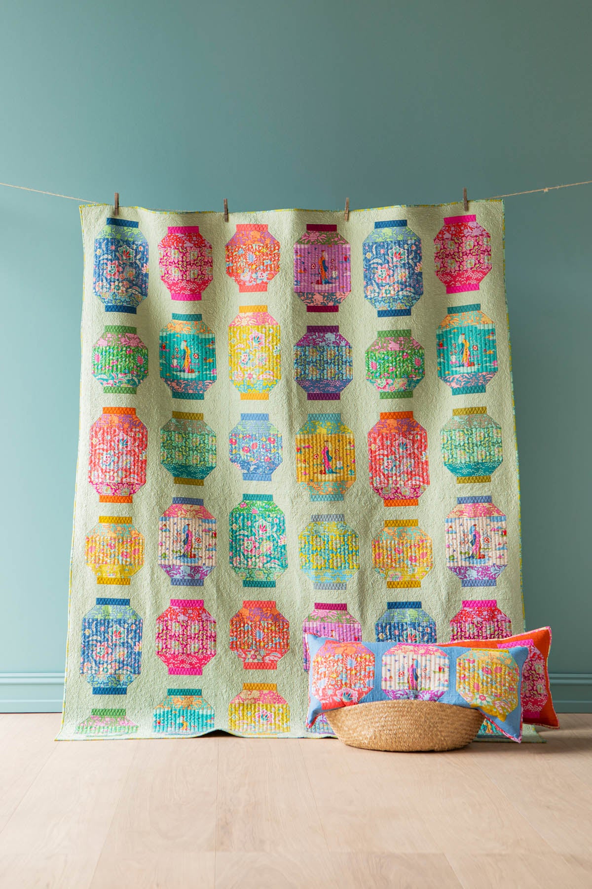 A colorful patchwork quilt featuring repeating geometric lantern-like patterns in pink, blue, green, yellow, and orange on a pale mint background, hanging from a string with wooden clothespins against a teal wall. A woven basket and matching accent pillows sit on the floor below.
Photo by Tildafabrics.com [additional_image_link]