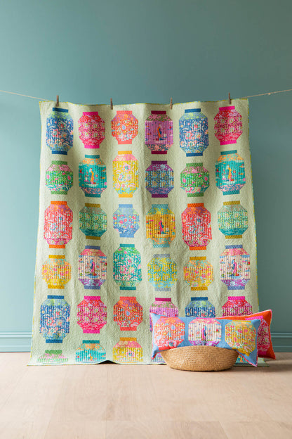 A colorful patchwork quilt featuring repeating geometric lantern-like patterns in pink, blue, green, yellow, and orange on a pale mint background, hanging from a string with wooden clothespins against a teal wall. A woven basket and matching accent pillows sit on the floor below.
Photo by Tildafabrics.com [additional_image_link]