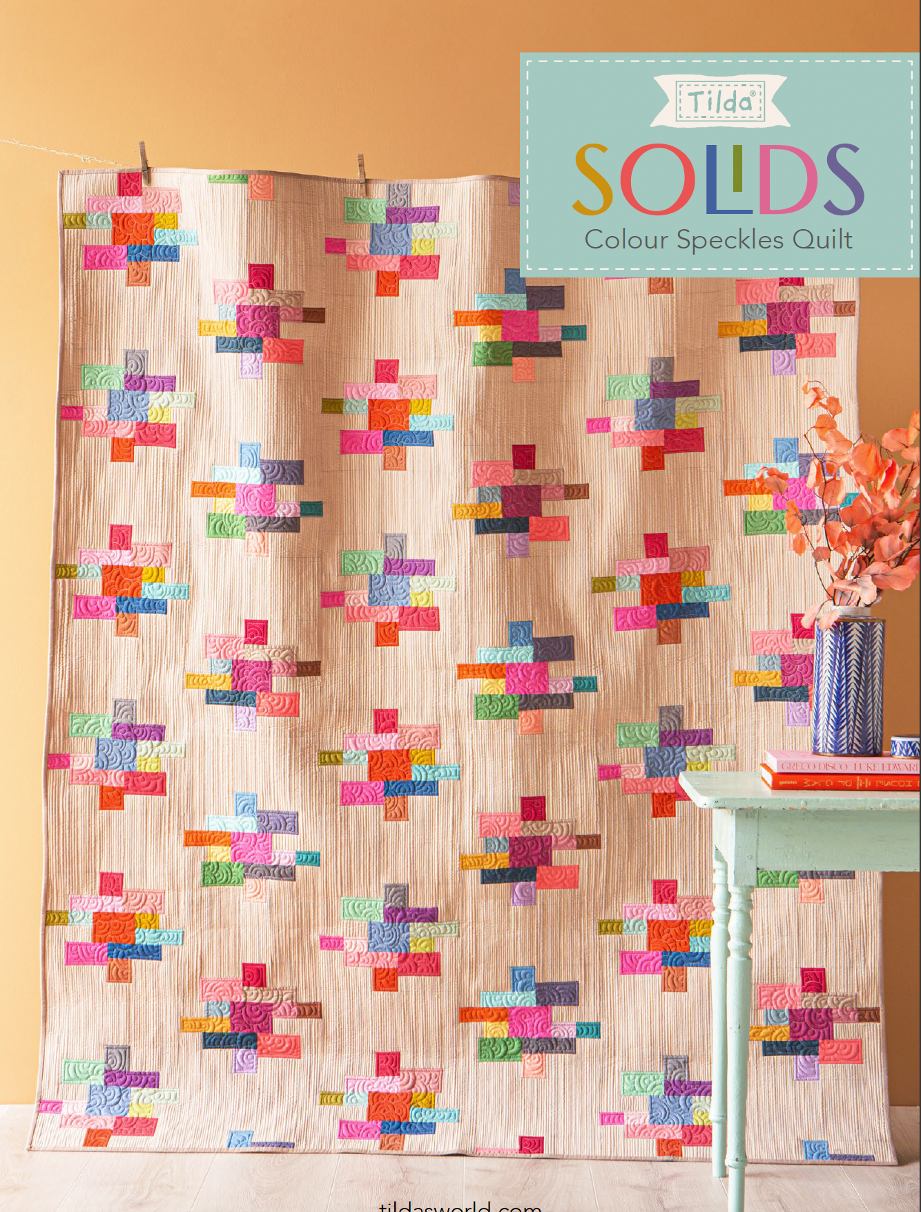A quilted textile panel or curtain featuring a repeating colorful speckled cross pattern on a beige striped background, shown hanging in an indoor setting with a small table and flowers beside it. A teal label in the top right reads 'Tilda SOLDS Colour Speckles Quilt'.
Photo by Tildafabrics.com [image_link]