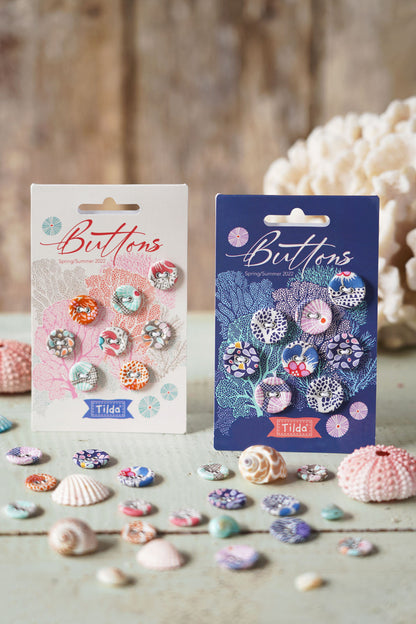 Two packages of Tilde buttons displayed on a rustic wooden surface with seashells and coral props. One package is white with pink floral artwork and shows multicolor patterned buttons, and the other is dark blue with teal floral artwork and shows purple-toned patterned buttons.
Photo by Tildafabrics.com [additional_image_link]