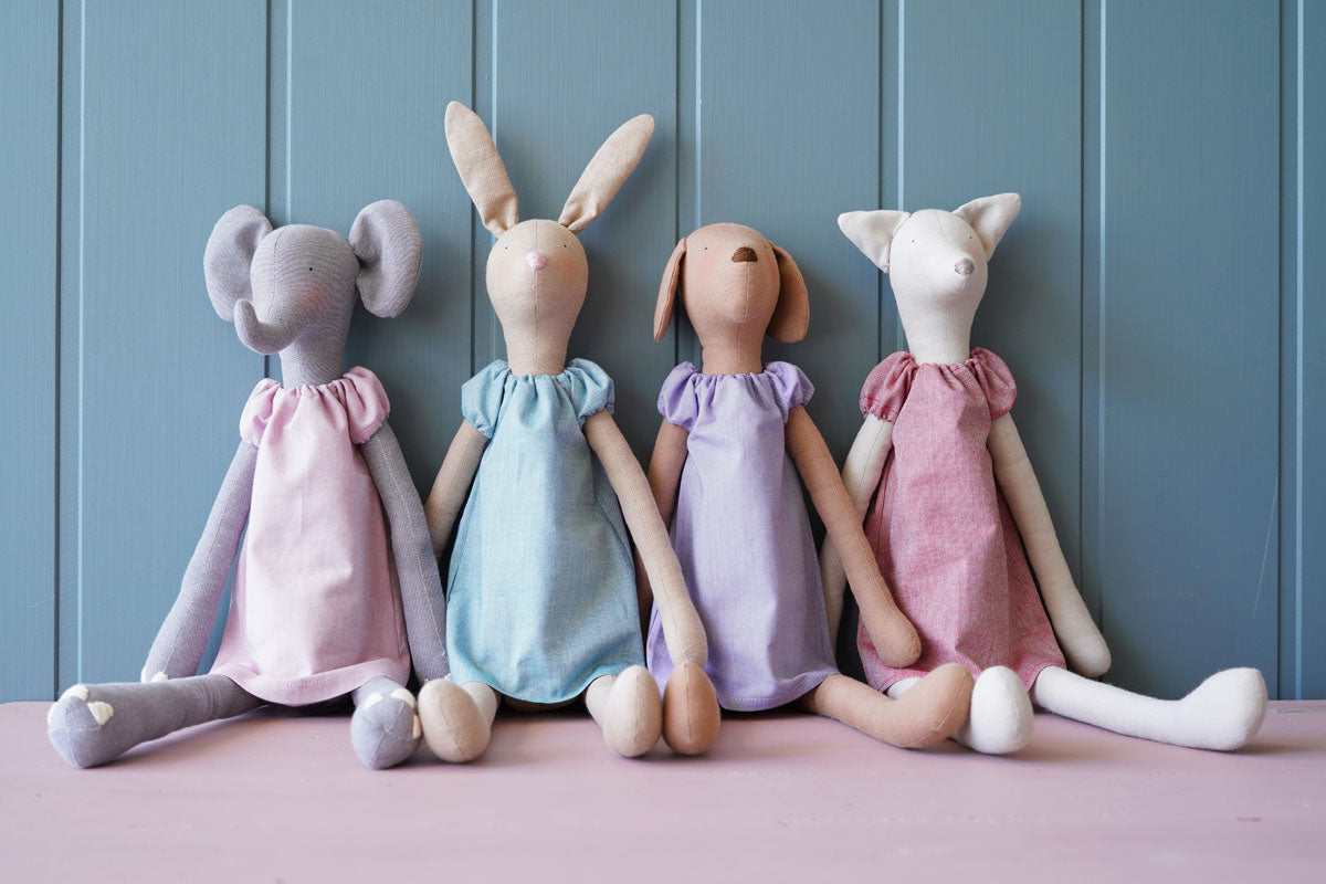 Four handmade fabric animal dolls sitting side by side against a blue paneled background. The dolls resemble a mouse, a rabbit, a dog, and a cat, each wearing a different pastel-colored dress in pink, light blue, lavender, and dusty pink.
Photo by Tildafabrics.com [additional_image_link]