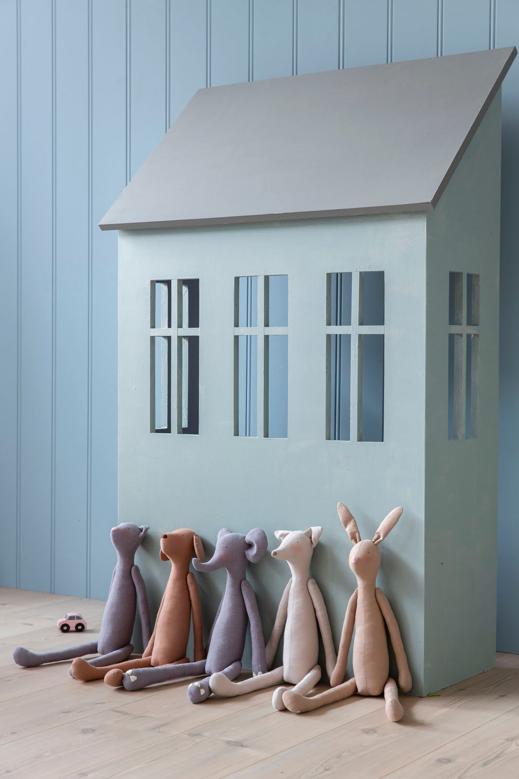 A large pastel house-shaped dollhouse or playhouse with three window cutouts, set against a light blue paneled wall and wood floor. Five soft stuffed animal figures in muted neutral colors sit along the base of the house.
Photo by Tildafabrics.com [additional_image_link]