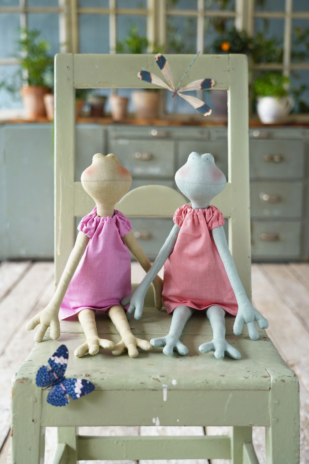 Two handmade fabric frog dolls wearing pink dresses sit on a distressed sage green wooden chair in a rustic room setting.
Photo by Tildafabrics.com [additional_image_link]
