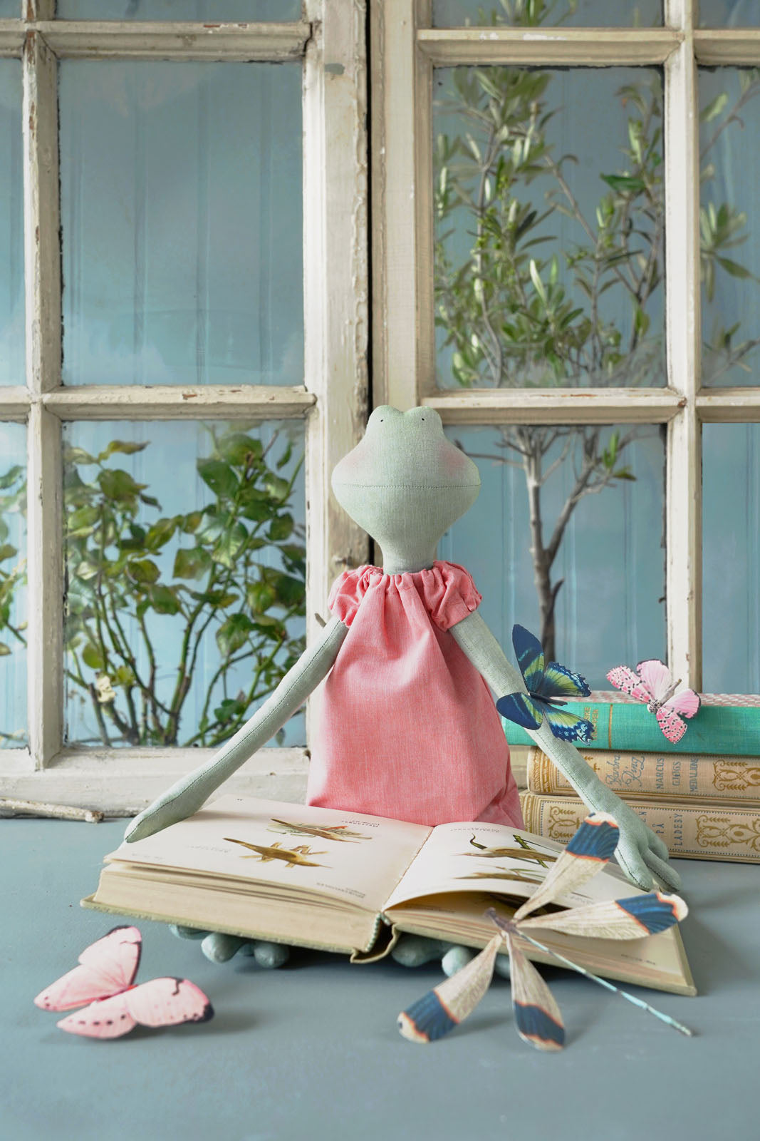 A whimsical handmade doll with long pale green arms and a pink dress sits on an open book in front of a vintage-style window. The scene includes pink butterflies, a small bird figurine, and stacked books on a soft blue-gray surface.
Photo by Tildafabrics.com [additional_image_link]