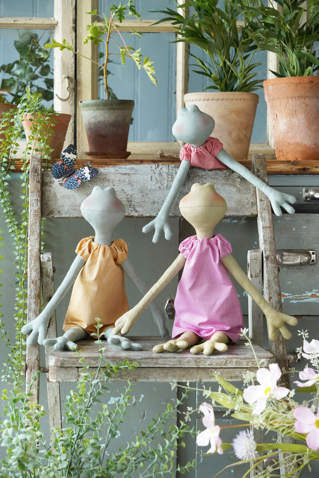 Three fabric doll figurines with long pale blue arms and legs, posed on a rustic wooden shelf among potted plants and greenery.
Photo by Tildafabrics.com [additional_image_link]